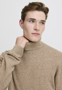 Maglione beige a collo alto in morbido materiale lavorato a maglia testurizzato con polsini e collo a coste, caratterizzato da un design classico aderente.