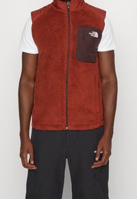 Chaleco de forro polar rojo con cuello alto, cierre completo y bolsillo en el pecho marrón. Presenta una superficie texturizada y detalles en colores contrastantes.
