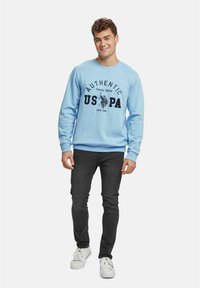 Lyseblå genser med rund hals som har trykket teksten "AUTHENTIC USPA" og logo, kombinert med mørkejeans og hvite sneakers.