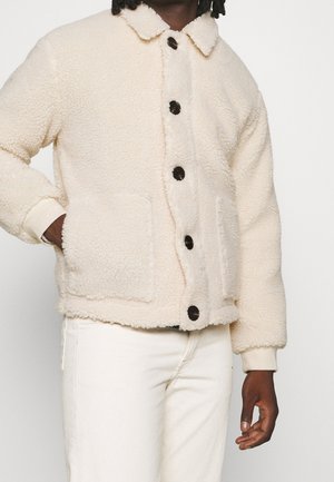 Personne portant une veste polaire crème avec de gros boutons noirs et des poches plaquées, associée à un pantalon blanc, sur un fond uni.