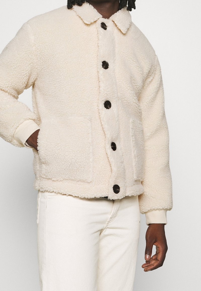 Personne portant une veste polaire crème avec de gros boutons noirs et des poches plaquées, associée à un pantalon blanc, sur un fond uni.