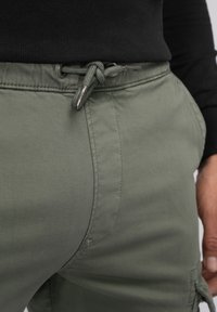 Olijfgroene broek van zachte stof, met een koord in de taille met metalen accenten en zijzakken, met een gladde textuur.