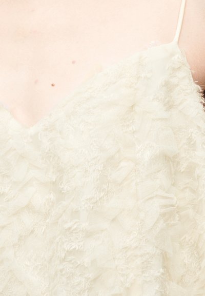 Επάνω μέρος από ελεφαντόδοντο ύφασμα με λαιμόκοψη σε σχήμα V, λεπτή υφή με βολάν και λεπτές τιράντες, με απαλό, διαφανές σχέδιο και χωρίς ορατά μεταλλικά στοιχεία.