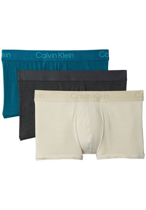 Muskelbepackter Mann, barfuß stehend vor neutralem Hintergrund, trägt türkisfarbene Calvin Klein Boxerbriefs mit den Händen in der Hüfte.
