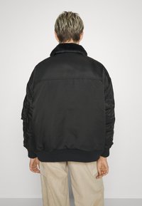 Sort bomberjakke med en fluffy krave og elastiske manchetter, der har en glat tekstur, struktureret form og samlede ærmer.