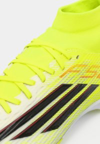 Φωτεινό κίτρινο ποδοσφαιρικό παπούτσι με υφή στην επιφάνεια, λεπτό προφίλ, μαύρες και κόκκινες λεπτομέρειες, καθώς και έναν γιακά και σύστημα κορδονιών που μοιάζουν με κάλτσα.