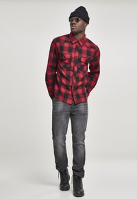 Rode en zwarte geblokte overhemdd met knopen aan de voorkant en twee borstzakken, gecombineerd met grijze jeans en zwarte veterschoenen. Accessoires zijn een beanie en een zonnebril.