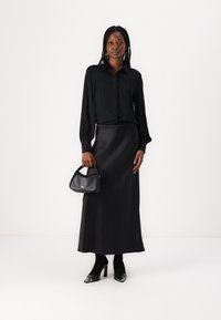 Black pleated blouse with long sleeves, paired with a black satin maxi skirt. Accessories include a small black handbag and heeled shoes.