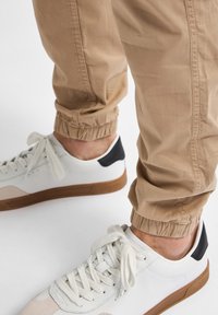 Lichtbeige joggers met elastische cuffs, gecombineerd met witte sneakers met een zwarte hakaccent en een tan kleurige zool. Gladde stoftextuur.
