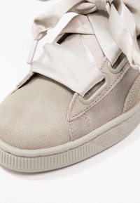 Zapatilla de deporte gris claro con parte superior de tejido texturizado, detalles en ante y cordones de cintas de satén. Suela de goma con un detalle estampado.