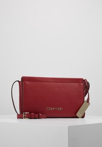 Czerwona skórzana torebka na ramię Calvin Klein z regulowanym paskiem, złotą plakietką z logo oraz dołączonym złotym prostokątnym tagiem na białej powierzchni.