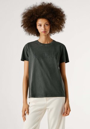 Camiseta de algodón verde oscuro con mangas cortas, cuello redondo y un sutil diseño de texto perforado en el frente, combinada con pantalones de color claro.