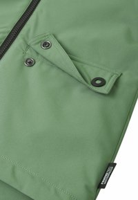 Dettaglio di un giubbotto impermeabile verde con una texture liscia, dotato di zip e tasca con scatto. Include un'etichetta nera con la scritta "reinotec impermeabile."