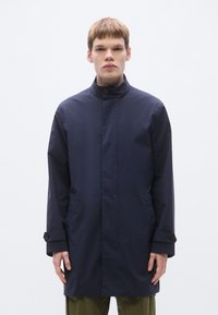 Tumesinine veekindel mantel kõrge kraega, ees nööbitav ja küljetaskutega; sileda tekstuuri ja minimalistliku disainiga.