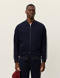 Ναυτικό μπλε φούτερ μπόμπερ με φερμουάρ, λευκές λωρίδες στο πλάι, ριμπ μανσέτες και ομαλή υφή. Δείχνεται με ένα ταιριαστό σύνολο και κόκκινο αξεσουάρ.