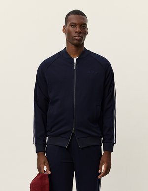 Jachetă bomber albastru închis cu fermoar, cu dungi laterale albe, manșoane ribbed și o textură fină. Prezentată cu o ținută asortată și un accesoriu roșu.