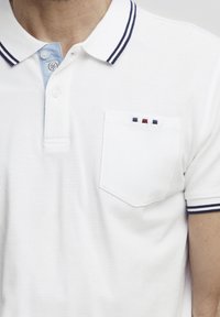 Hvit polo skjorte laget av strukturert stoff, med en blå krage med marineblå striper og en brystlomme med tre fargede detaljer (rød, blå, hvit).