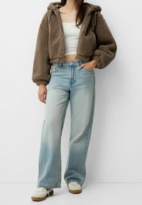 Bruine, korte, pluizige hoodie met capuchon, gecombineerd met lichtblauwe, wijd uitlopende jeans. Witte sneakers met beige accenten maken de look af.