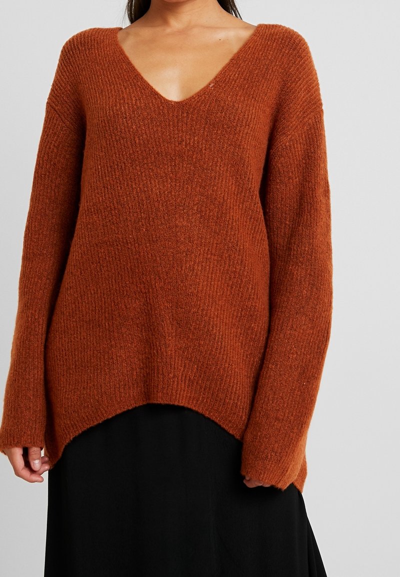 Femme portant un pull en tricot ample couleur rouille avec col en V et manches longues, assorti à une jupe noire, sur un fond uni.