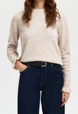 Femme portant un pull beige à manches longues rentré dans un jean bleu foncé avec une ceinture noire, une main dans la poche, cheveux bouclés visibles.