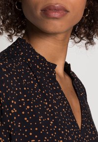 Blusa di tessuto nero con punti arancioni sparsi, colletto arricciato e design a V. Presenta una texture liscia e un leggero lucido.