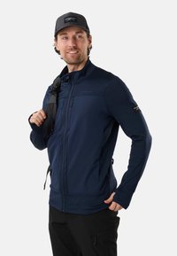 Homme portant une veste zippée bleu marine et une casquette noire, avec une sangle de sac à dos sur une épaule, regardant sur le côté et souriant.