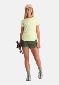 Light green, short-sleeve athletic shirt, paired with dark green shorts. Gray hiking shoes and a beige cap. Holding hiking poles.