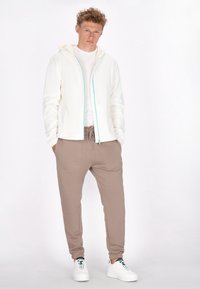 Lichtgewicht crème hoodie-jacket met turquoise rits, gecombineerd met beige joggers met zijzakken. Afgerond met witte sneakers.