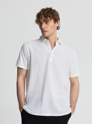 Uomo con capelli ricci che indossa una polo bianca a maniche corte con texture e pantaloni neri, in piedi con le mani in tasca davanti a uno sfondo semplice.