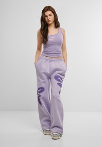 Ljuslila linne med en liten rosa logotyp, tillsammans med matchande lösa sweatpants som har mörklila abstrakta mönster.