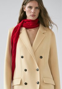 Beiger zweireihiger Blazer aus Wollmischung mit schwarzen Knöpfen, kombiniert mit einem strukturierten roten Zopfmuster-Strickschal, der um den Hals gewickelt ist.