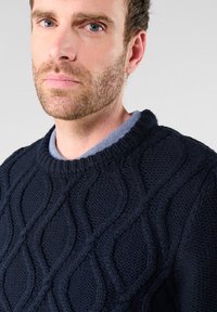 Gestreifter marineblauer Pullover mit einem strukturierten Wellenmuster, rundem Ausschnitt, über einem hellblauen Hemd mit Kragen getragen, aus dickem Stoff.