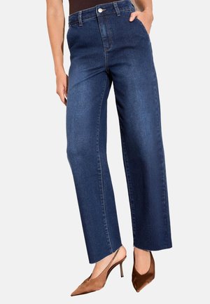 Kobieta ubrana w ciemnoniebieskie dżinsy o wysokim stanie i szerokich nogawkach oraz brązowe buty na obcasie typu slingback ze spiczastym noskiem, stoi z jedną ręką w kieszeni.