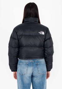 Persona con lunghi capelli scuri che indossa un giubbotto invernale nero e jeans azzurri chiari, girata di spalle, con il logo The North Face sul giubbotto.