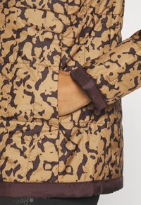 Main portant une veste matelassée imprimé léopard avec un poignet marron, main partiellement dans une poche avant zippée.