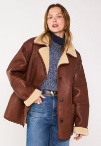 Veste en cuir marron avec col et poignets en peau de mouton, dotée d'une fermeture à boutons et de grandes poches latérales, portée par-dessus un pull en tricot bleu et un jean.
