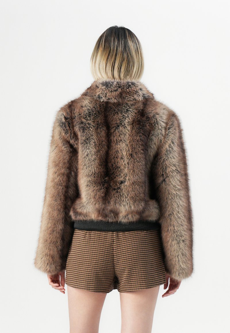 Osoba z krótkimi włosami ombre, ubrana w brązową kurtkę z futra sztucznego i brązowe szorty w wzór houndstooth, stoi tyłem na białym tle.