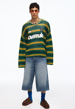 Jonge man die een groene en geel gestreepte gaas trui draagt met de tekst "avenue", baggy spijkershort, blauwe sokken, bruine schoenen en rode zonnebril.