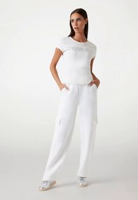 T-shirt en coton blanc avec logo "GUESS" en finition métallique, associé à un pantalon cargo blanc ample avec poches latérales. Baskets argentées.