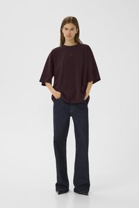 Ung kvinne står med hendene i lommene, iført en løs mørk lilla oversized T-skjorte og mørk blå vide bukser mot en ensfarget bakgrunn.