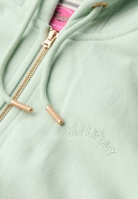 Felpa con zip di colore verde menta, realizzata in tessuto morbido, dotata di zip in metallo dorato e logo ricamato "SUPERDRY".