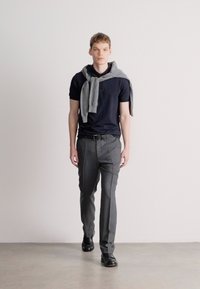 Μαρινί πουκάμισο τύπου πόλο, γκρι πουλόβερ ριγμένο στους ώμους, γκρι παντελόνι, μαύρα παπούτσια, στενός σχεδιασμός, μινιμαλιστική εμφάνιση, όρθιος σε ουδέτερο φόντο.