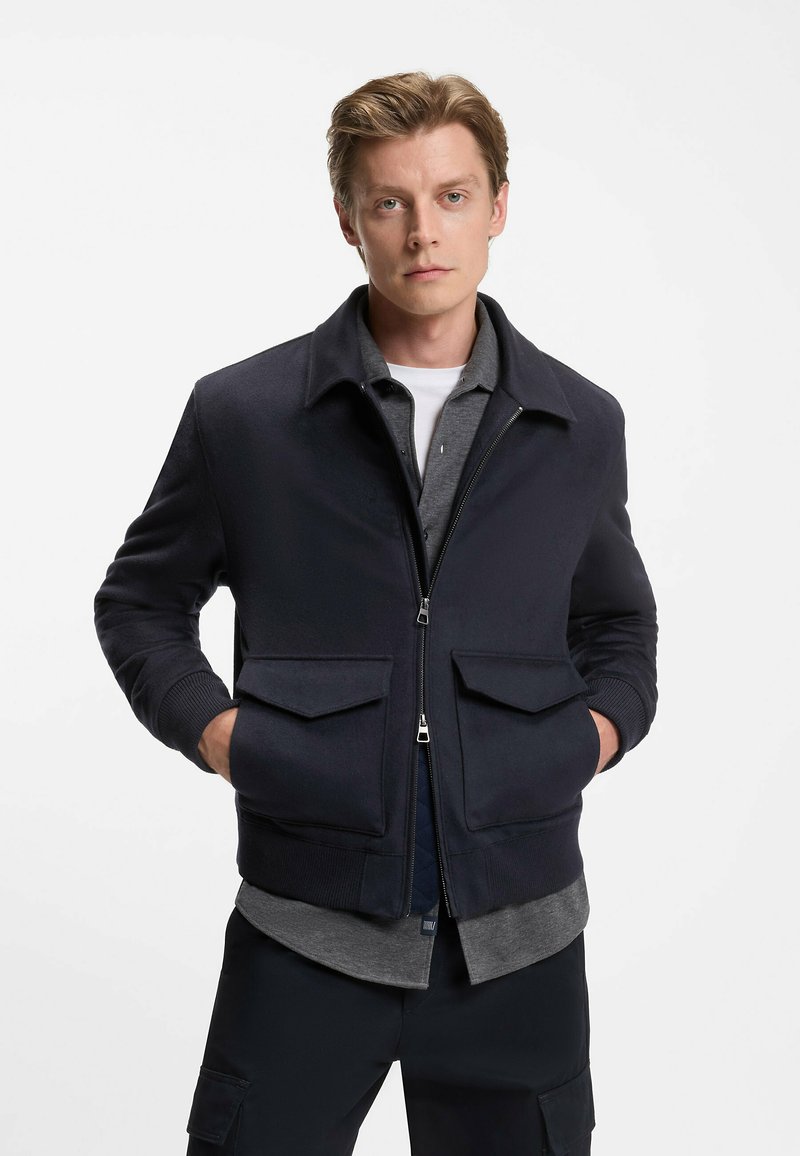 Navy bomber jacket with a zip front, two front pockets, elastic cuffs, and a collared neckline, layered over a gray shirt.