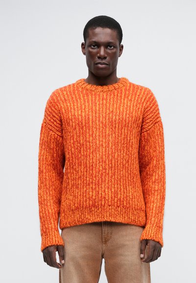 Pulover chunky portocalie din tricot, cu un model texturat de dungi, guler rotund și umeri căzuți, asortat cu pantaloni bej deschis.