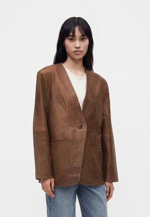 Ung kvinde med langt mørkt hår iført en brun læderblazer over en hvid top og blå jeans, stående mod en ensfarvet lys baggrund.