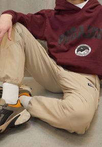 Osoba v béžových kalhotách, bílých ponožkách a černých a oranžových teniskách, sedící na podlaze, oblečená v bordovém svetru s nápisem "PEGADOR" a logem hory.