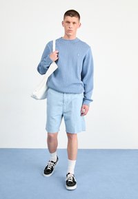 Suéter de punto azul claro con textura acanalada, combinado con pantalones cortos azul claro, zapatillas negras y un bolso tote blanco.