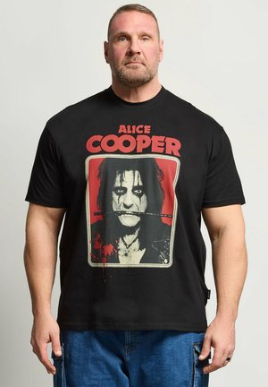 Mann mit kurzen Haaren und Bart, der ein schwarzes Alice Cooper T-Shirt mit dem Gesicht des Sängers und rotem Text trägt, kombiniert mit blauen Jeans.