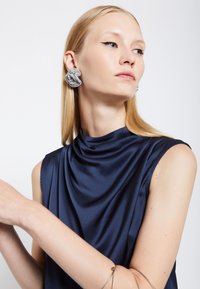 Námořnická saténová blůzka s šatkovým výstřihem a bezramínkovým designem, doplněná texturovanými stříbrnými náušnicemi připomínajícími propletené tkaniny.