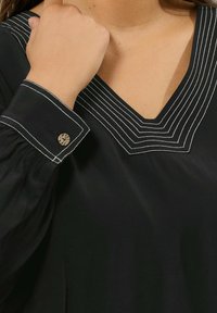 Blusa nera con scollo a V caratterizzata da dettagli in cucitura bianca e maniche puff. Il polsino ha un accento decorativo con bottone metallico.